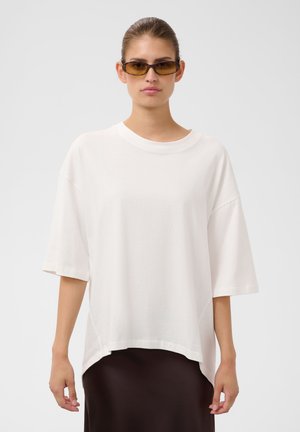 Femme de face portant un t-shirt blanc oversize à manches courtes, une jupe marron et de petites lunettes de soleil rectangulaires teintées marron sur fond gris uni.
