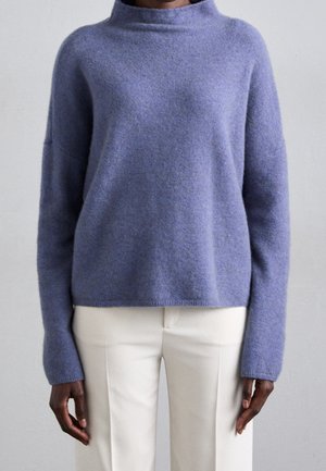 Osoba ubrana w lawendowy, długorękawowy sweter o miękkiej fakturze i wysokim dekolcie oraz białe spodnie na jednolitym tle.