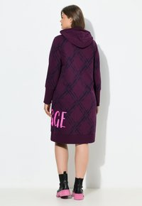 Donna che indossa un vestito con cappuccio viola, decorato con un motivo a quadri scuri e testo rosa vicino all'orlo, abbinato a stivali neri alla caviglia con suole rosa, in posizione laterale.