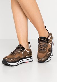 Zapatillas marrones con material de malla y ante, detalles metálicos, laterales con patrones y una suela gruesa blanca y negra. Logo en la lengüeta.