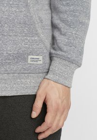 Sivý sweatshirt s textúrovanou látkou, rebrovanými manžetami a malou nálepkou s nápisom "BLEND" na leme. Časť ruky je viditeľná, položená na tmavej látke.