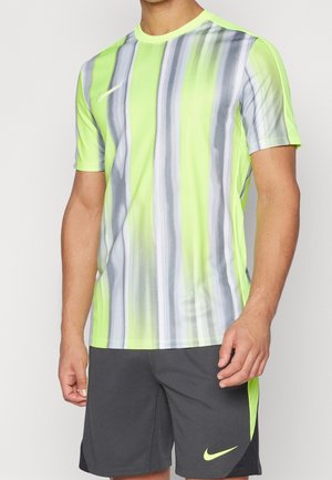 Mężczyzna ubrany w neonowo-żółtą i szarą sportową koszulkę z krótkim rękawem w paski oraz ciemnoszare spodenki z neonowo-żółtymi logo Nike, stojący na jednolitym tle.