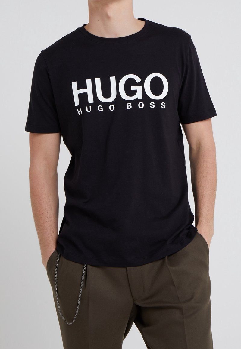 Zwart katoenen T-shirt met in het wit de opvallende tekst "HUGO HUGO BOSS" op de voorkant. Korte mouwen en een ronde hals, gecombineerd met olijfgroene broek.