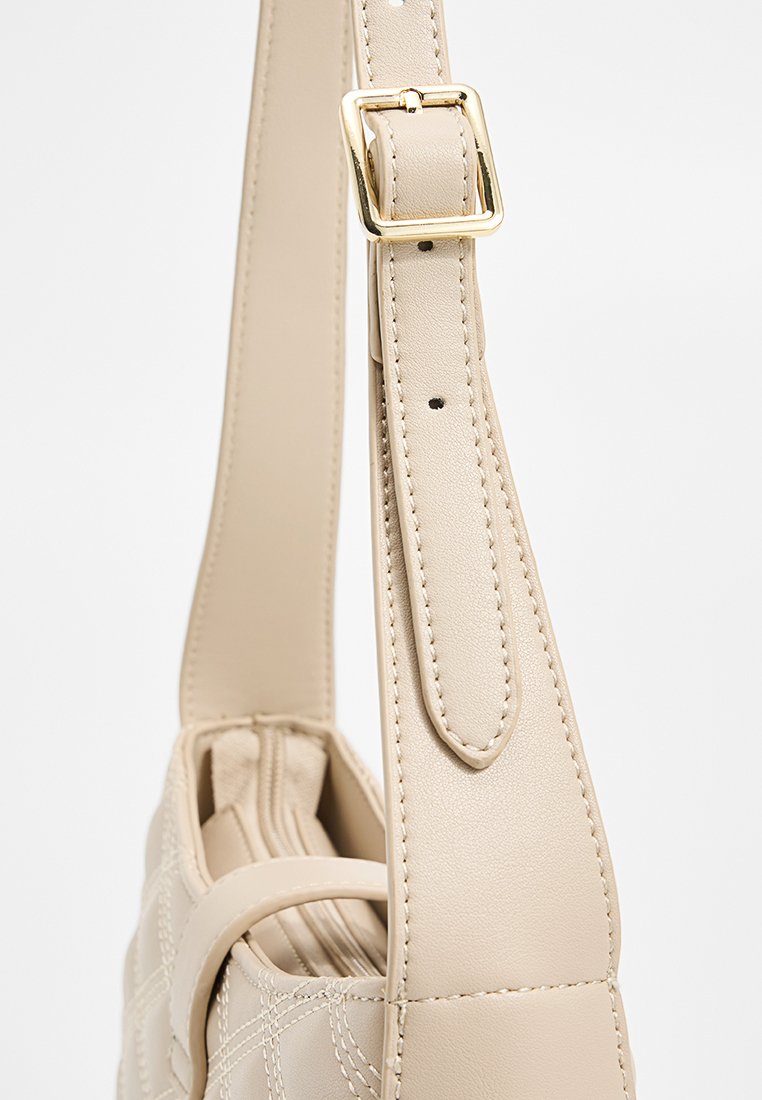 Beige Schultertasche mit strukturierter Oberfläche, ausgestattet mit einem breiten Riemen und einer goldenen Schnalle, die durch gestickte Details hervorgehoben wird.