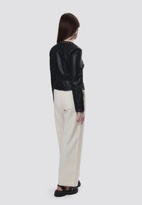 Giacca di pelle nera con un design aderente, caratterizzata da borchie lungo il collo, abbinata a pantaloni a gamba larga color crema.