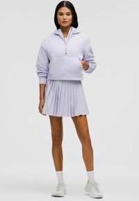 Sudadera recortada lila con bolsillo frontal y cremallera plateada, combinada con una falda plisada a juego. La modelo lleva zapatillas y calcetines blancos.
