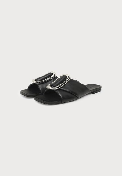 Paire de sandales plates noires avec larges brides en cuir et grandes boucles ovales argentées sur fond blanc uni.