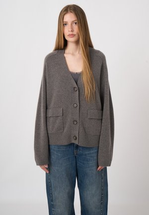 Young woman with long straight hair wears oversized gray buttoned cardigan and wide-leg blue jeans against plain background.