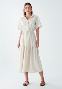 Šviesiai smėlinis mygtukinis suknelė su trumpomis rankovėmis, su raišteliu juosmenyje ir daugiasluoksne sijonu. Lygus audinys, vidutinio ilgio ir be matomų raštų.