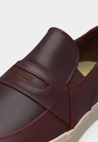 Κοντινή λήψη ενός καφέ δερμάτινου παπουτσιού χωρίς κορδόνια, με ελαστική πλαϊνή πλευρά, διακοσμητικό λουράκι και μπεζ καουτσούκ σόλα.