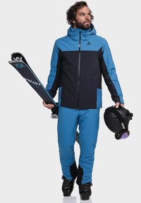 Blaue und schwarze Skijacke mit Kapuze, kombiniert mit passenden blauen Hose. Hält Skier und einen schwarzen Helm mit einem reflektierenden Visier.