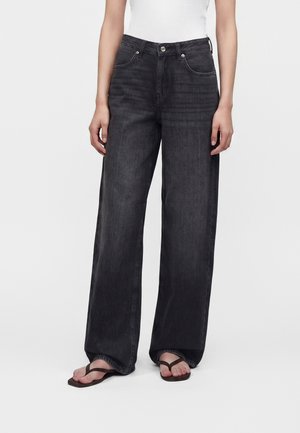 Person iført sorte vidde bukser med høj talje, hvid top og brune flip-flops, stående mod en ensfarvet baggrund.