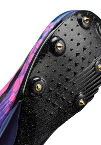 Gros plan sur la semelle d'une chaussure avec des crampons cloutés noirs, quatre pointes dorées proéminentes, et un bord supérieur rose-violet avec le texte "FLYPLATE".