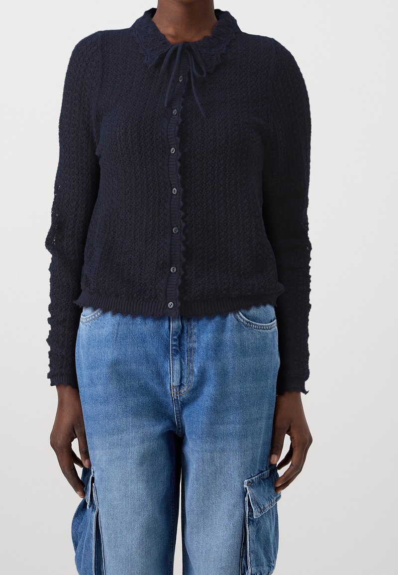 Personne portant un cardigan texturé bleu marine boutonné avec un col noué et un pantalon cargo en denim bleu clair.