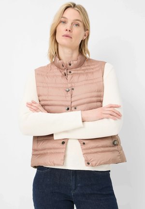 Frau mit verschränkten Armen, die eine hellbraune gesteppte Weste über einem weißen Langarmshirt und dunkelblaue Jeans vor weißem Hintergrund trägt.