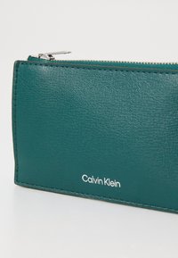 Žalias odinis piniginė su tekstūriniu paviršiumi, turintis sidabrinį užtrauktuką ir baltai spausdintu logotipu „Calvin Klein“ šone.