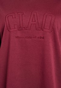 T-shirt bourgogne avec "CIAO" brodé en évidence dans une teinte plus foncée et "état d'esprit Milano" en dessous dans un fil assorti, texture lisse.
