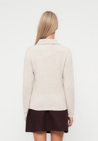 Beige neulepusero, jossa on kauluksellinen pääntie, pitkät hihat, kirjailut hihansuut ja kapea helma, yhdistettynä tummiin hameeseen. Takakatsaus.