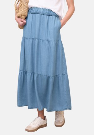 Kvinde iført en lyseblå maxinederdel i lag med elastisk talje, hvide sneakers og med en flettet stråtaske, med den ene hånd i lommen.