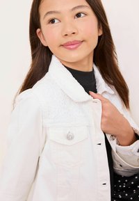 Valkoinen farkkutakki, jossa kauluksessa pitsikoristeita, hopeiset napit ja teksturoitu pinta; päällä musta pilkullinen paita.