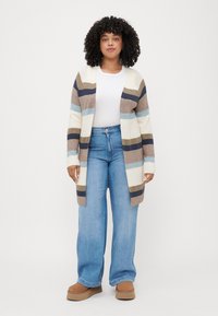 Striped cardigan in cream, brown, blue, and navy; open front design; soft, knitted texture; paired with light blue wide-leg jeans and brown boots.