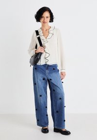 White long-sleeve blouse with black ruffle trim, paired with loose blue jeans featuring black floral appliqués, and black slip-on shoes.