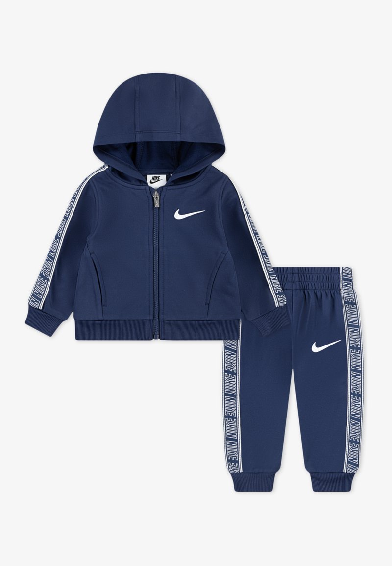 Súprava teplákov v námorníckej modrej farbe obsahujúca zipsovú mikinu a nohavice, pričom oboje je zdobené bielym logom Nike. Kontrastné pruhy na rukávoch a nohaviciach. Mäkký materiál.