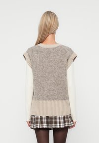 Beige strikket vest med korte ermer, med en teksturert rygg og ribbestrikket kant, lagt over en hvit langermet skjorte og rutete skjørt.
