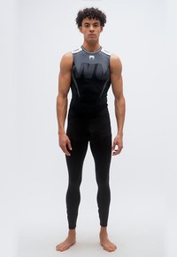 Νεαρός άνδρας που στέκεται ξυπόλητος, φορώντας αθλητικό τοπ χωρίς μανίκια σε μαύρο και γκρι και μαύρο κολάν, μπροστά σε λευκό φόντο