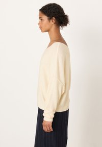 Kvinna i krämfärgad off-shoulder fluffig tröja och marinblå randig kjol står i profil mot en enkel ljus bakgrund.