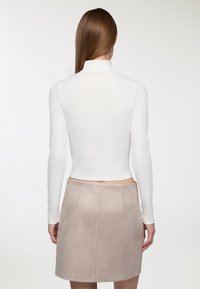 Kobieta z długimi prostymi włosami, ubrana w białą bluzkę z długim rękawem i beżową spódnicę z zamszu, stojąca tyłem.