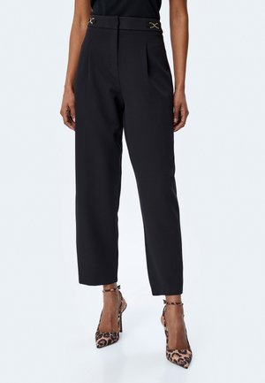 Hooggewaisted zwarte broek met plooien, met gouden accenten op de tailleband. Glad stof en een taps toelopende pasvorm, gecombineerd met schoenen met luipaardprint.