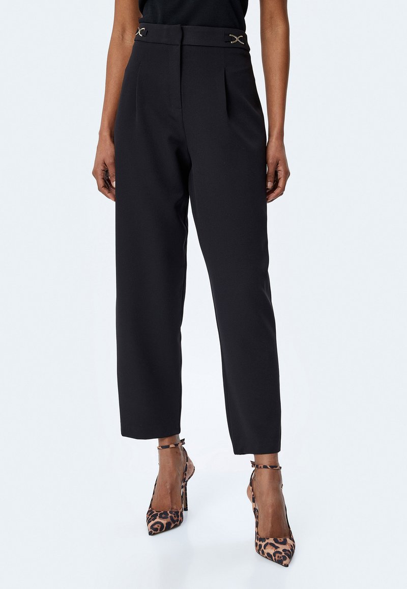 Czarne spodnie z wysokim stanem i plisami, z złotymi akcentami w talii. Gładki materiał i zwężany krój, w połączeniu z leopardowymi szpilkami.