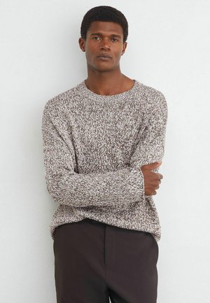 Mann mit kurzen lockigen Haaren, der einen braunen und weißen gestrickten Pullover und dunkle Hosen trägt, steht mit verschränkten Armen vor einer einfarbigen weißen Wand.