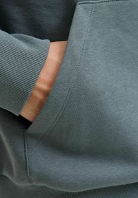 Sweatshirt vert menthe avec des poignets côtelés, poche kangourou et texture de tissu douce, avec des détails de couture visibles sur les coutures.