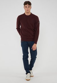 Maglione bordeaux a girocollo con maniche lunghe, polsini e orlo a coste; abbinato a jeans blu scuro e sneakers bianche con dettagli marroni.