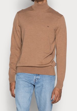 Brauner Strickpullover mit Rollkragen, langen Ärmeln und gerippten Bündchen, kombiniert mit hellblauen Jeans. Kleines Logo auf der Brust.
