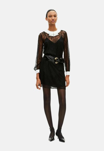 Robe en dentelle noire avec manches longues, col blanc, et une taille marquée soulignée par une ceinture noire. Collants noirs et chaussures vernies complètent la tenue.