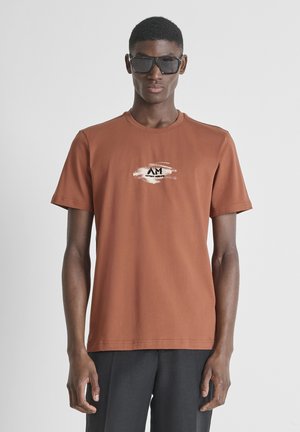 Muškarac koji nosi pretjerano velike crne sunčane naočale i hrđasto narančastu majicu s malim logotipom, stoji ispred jednobojne svijetle pozadine.