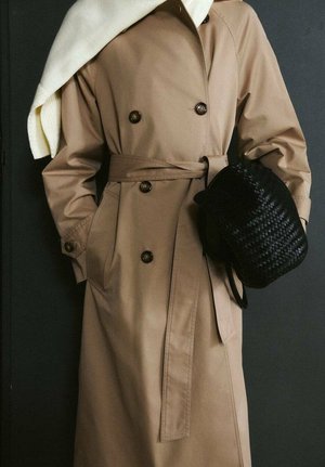 Kobieta w beżowym trenczu z paskiem, trzymająca czarną plecioną torebkę, z białym szalikiem zarzuconym na ramię.