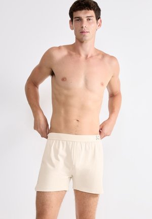 Beige Baumwollboxershorts mit einem elastischen Bund. Der Stoff ist glatt mit minimaler Textur und einem kleinen Logo am Bund.