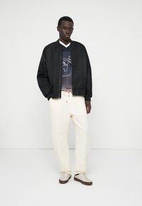 Veste pilote noire, t-shirt graphique blanc, pantalon crème et baskets beiges. Texture lisse, coupe décontractée et design minimaliste.