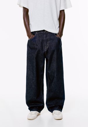Person mit lockeren, dunkelblauen Jeans, weißen Sneakers und einem weißen, oversized kurzärmeligen Hemd, die Hände in den Taschen vor einem weißen Hintergrund.