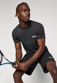 T-shirt de sport noir à manches courtes avec un design texturé et le logo "BOSS", associé à un short noir, tenant une raquette de tennis.