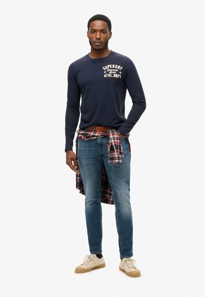 Marineblauw overhemd met lange mouwen met "SUPERDRY" logo, blauwe jeans en een rood geruit overhemd om de taille geknoopt. Beige sneakers maken de look compleet.