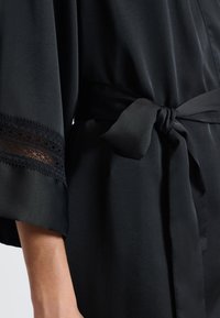 Raso nero con cintura annodata, maniche larghe e delicata finitura in pizzo ai polsini. Texture liscia con un design fluido.