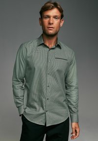 Green checkered long-sleeve shirt featuring a button-down collar, front pocket, and a smooth texture. Worn with dark pants.