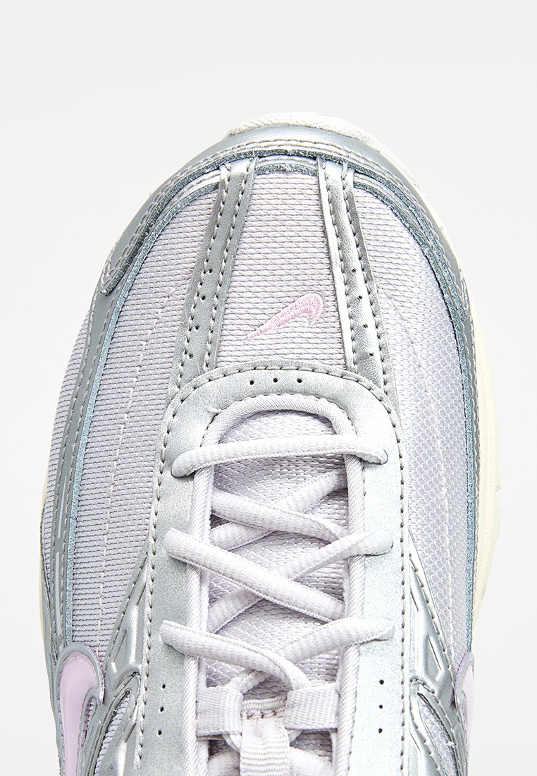 Zapatilla gris claro con acentos plateados, textura de malla y logo de Nike en rosa. Presenta punta redondeada, detalles de costura y cordones planos.