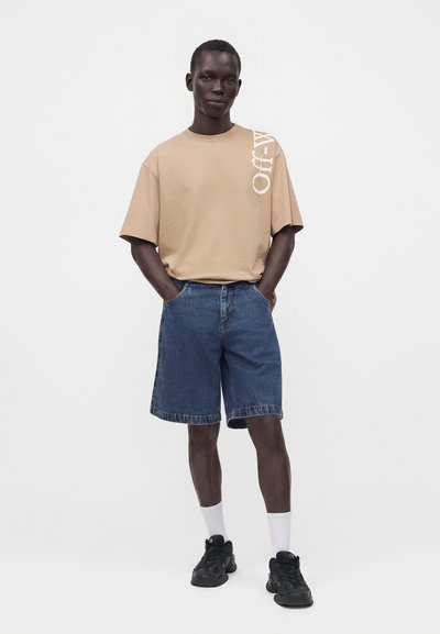 Homme portant un t-shirt beige oversize avec texte blanc, short en jean bleu, chaussettes blanches et baskets noires, debout les mains dans les poches.