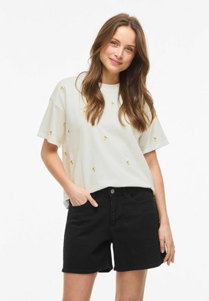 Jeune femme aux longs cheveux bruns portant un t-shirt blanc avec de petits imprimés de fruits jaunes et un short noir, souriante, une main dans la poche.